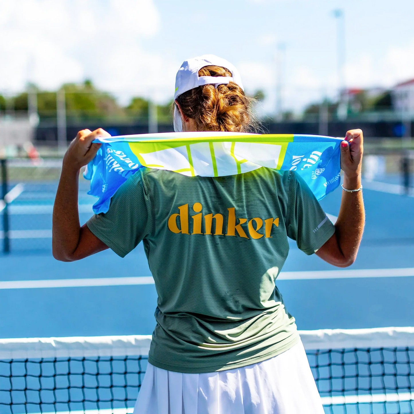 On a sunny blue court, a woman wears the Swinton Pickleball Women's Dinker LUX Performance Shirt in green with "dinker" in yellow, paired with a white skirt, and holds a colorful towel behind her neck as she gets ready to play.