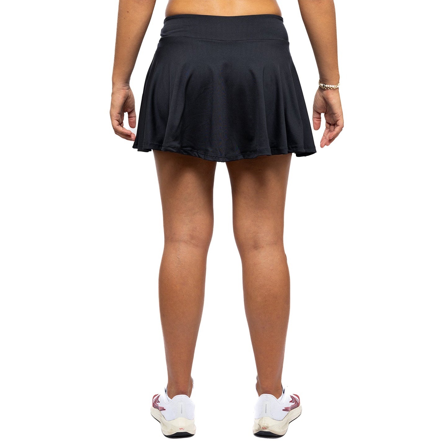 A woman faces away from the camera against a plain white background, modeling the Diadem Sports Women's Essential Club Skirt in black. Only her lower back, skirt, legs, and feet in white sneakers with red accents are visible.