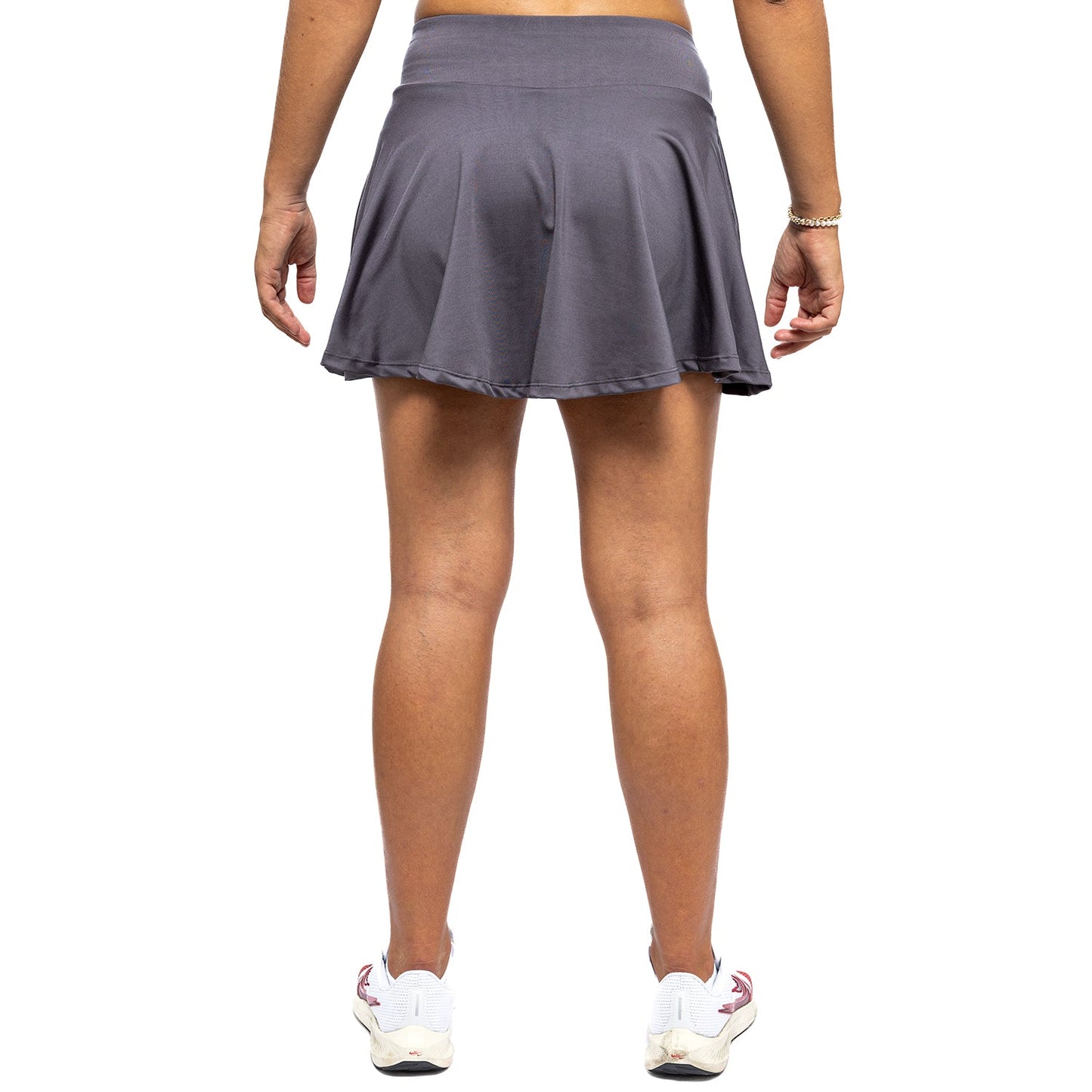 A woman is seen from the back wearing Diadem Sports' Women's Essential Club Skirt in gray, paired with white athletic shoes featuring maroon accents, standing relaxed against a plain white background.