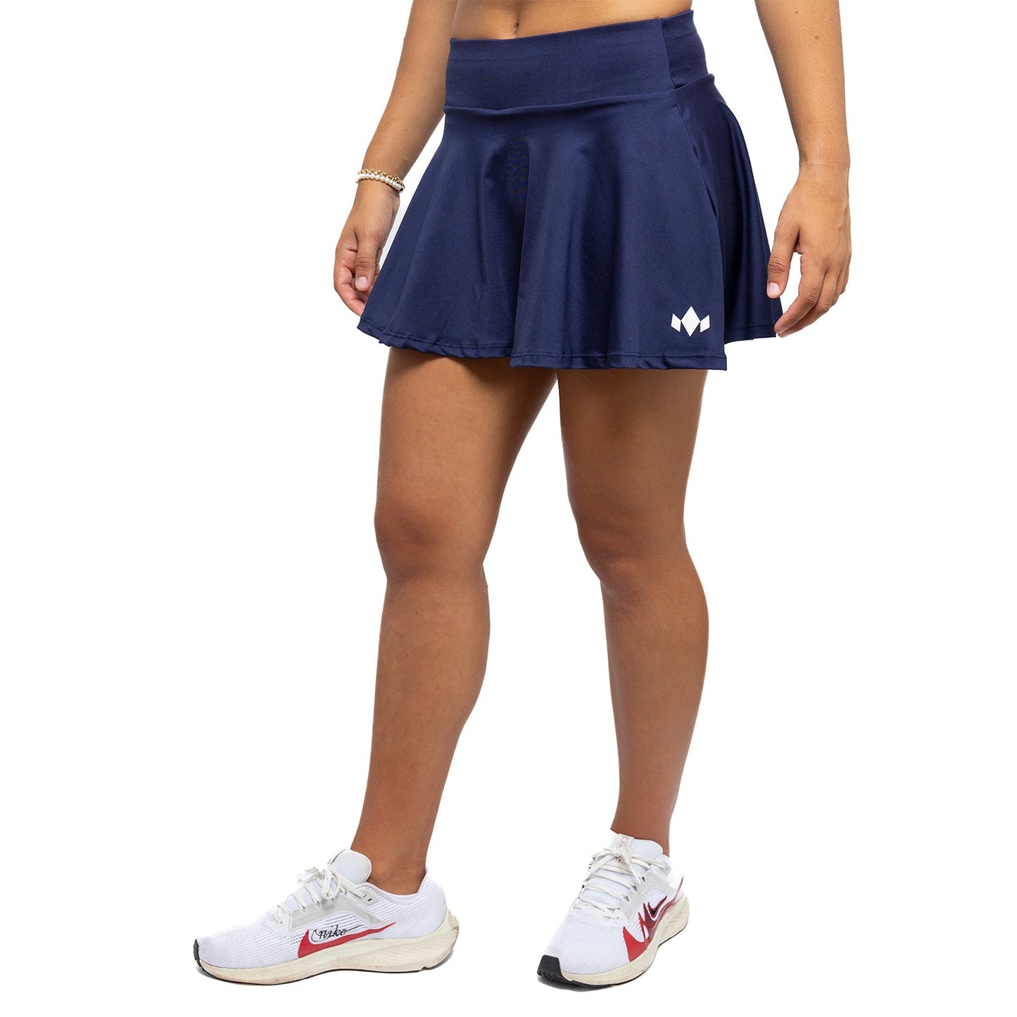 A person wears the Diadem Sports Women's Essential Club Skirt in navy blue, paired with white sneakers with red accents and a gold bracelet. Only their lower half appears against a plain white background.