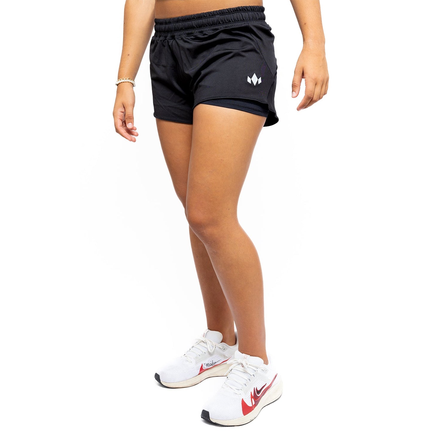 A person wears Diadem Sports Women's Essential Shorts featuring a small white geometric logo on the left leg, a bracelet on their right wrist, and white running shoes with red and black Nike branding, standing against a white background.