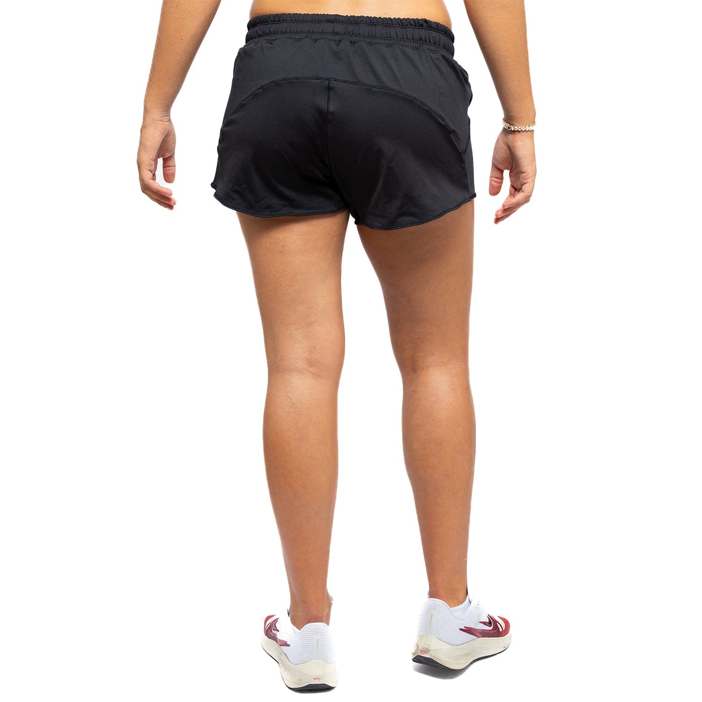 A person, shown from the waist down against a plain white background, wears Diadem Sports Women's Essential Shorts in black, paired with a gold bracelet and white sneakers featuring red and black accents.