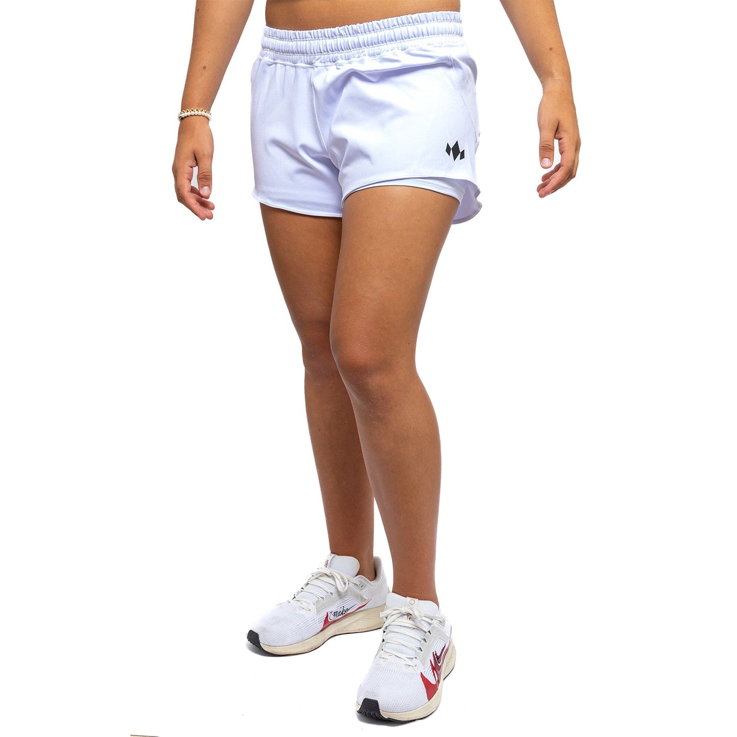 A woman stands against a white background, shown from shoulders down, wearing Diadem Sports Women's Essential Shorts with a small black logo on the left leg and white running shoes with red accents. Her arms are relaxed at her sides.