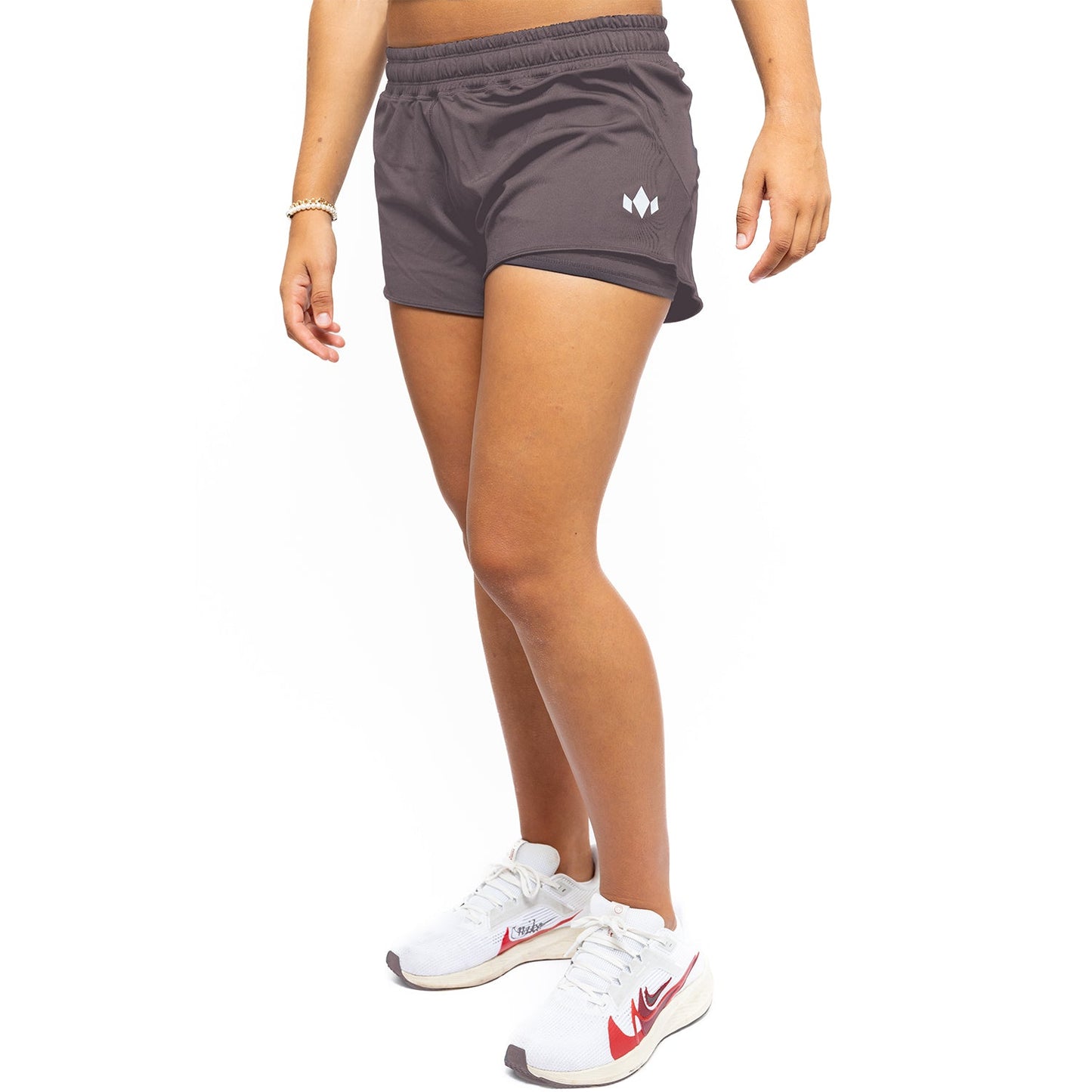 Only the lower body is visible against a white background, wearing Diadem Sports Women's Essential Shorts in charcoal gray with a white logo on the left and white Nike running shoes with red swooshes; hands rest at the sides.
