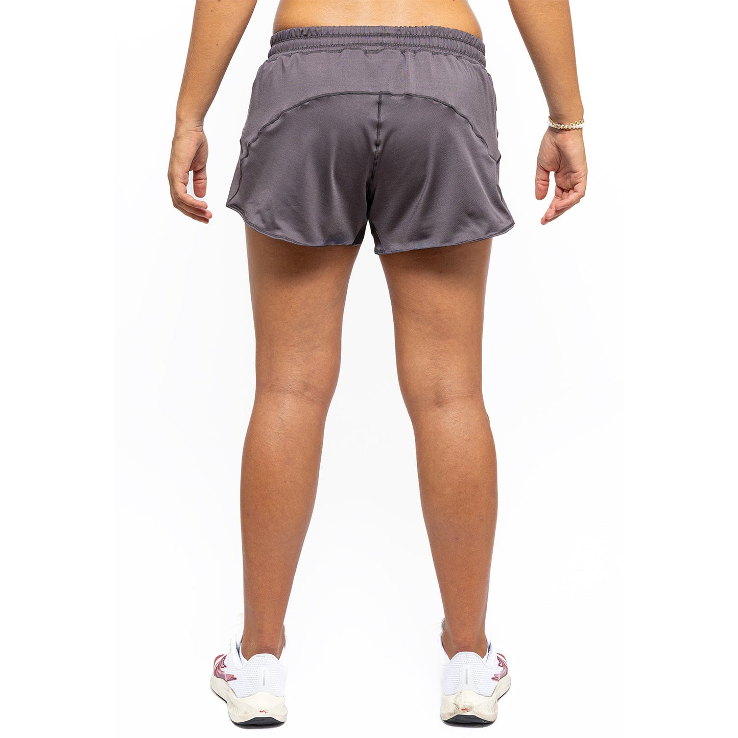A person’s lower body is shown from behind, wearing Diadem Sports Women's Essential Shorts and gray-white sneakers with light accents. Arms are relaxed at their sides against a plain white background.