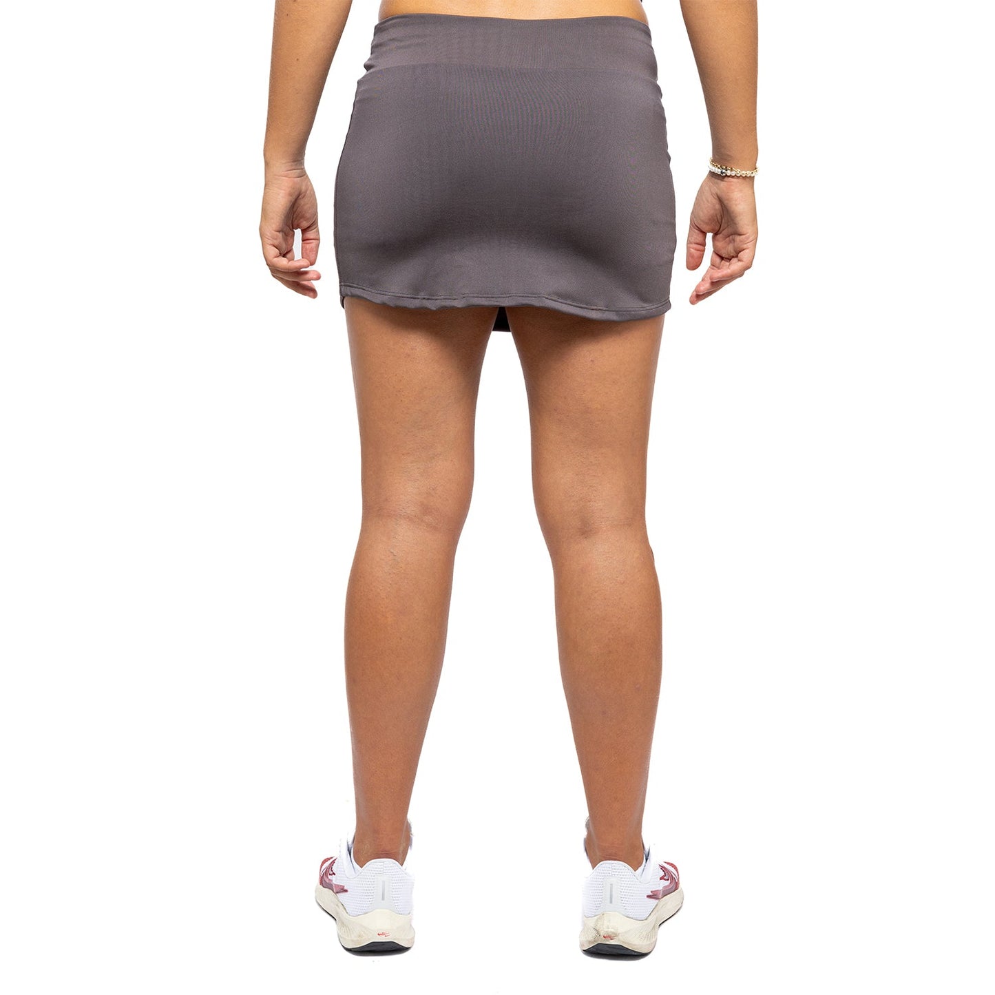 A person is shown from behind wearing the Diadem Sports Women's Essential Tennis Skirt, made of quick-dry fabric, and white athletic shoes with red accents. Only their lower back, legs, and shoes are visible; a bracelet adorns their right wrist.