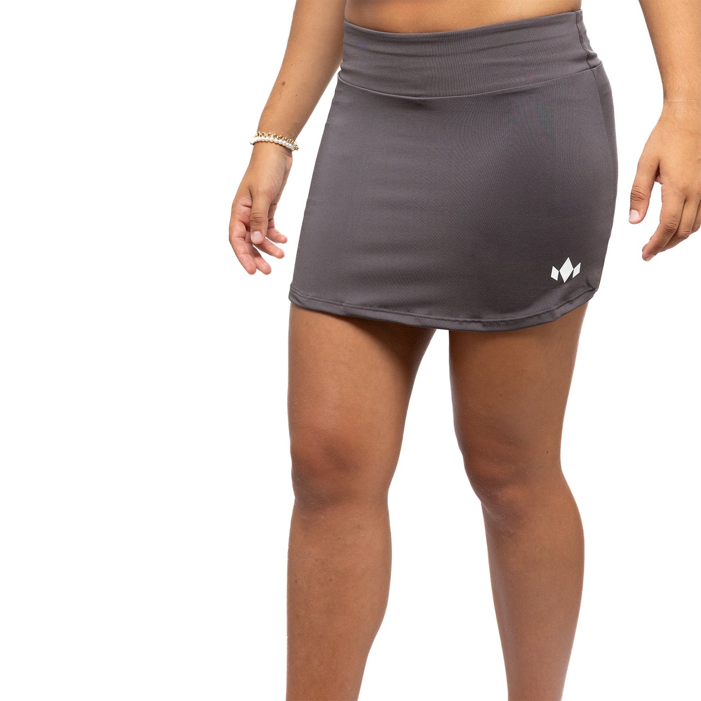 A person wears a dark gray Diadem Sports Women's Essential Tennis Skirt with a high waistband and small white geometric logo on the left. Made from fast-drying fabric, this athletic skirt provides comfort and performance compression. The background is plain white.
