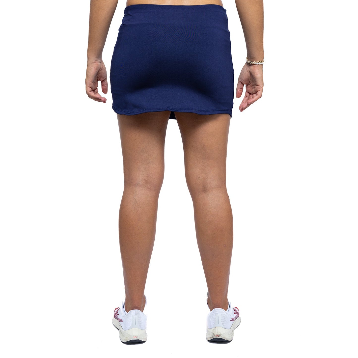 A person is seen from the back wearing Diadem Sports' Women's Essential Tennis Skirt in navy blue and white sneakers with red accents. Their arms are relaxed by their sides, a thin bracelet on the right wrist, against a plain white background.