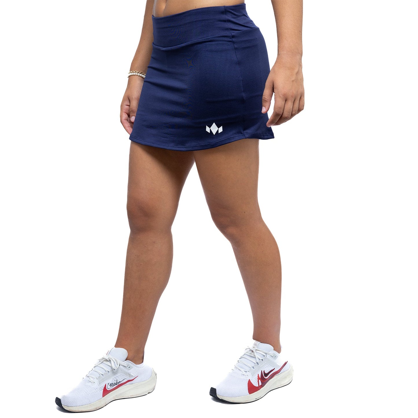 A person wears a Diadem Sports Women's Essential Tennis Skirt in navy blue, made from fast-drying fabric with a small white logo on the left, paired with white ankle socks and Nike running shoes. Upper body is partially out of frame.