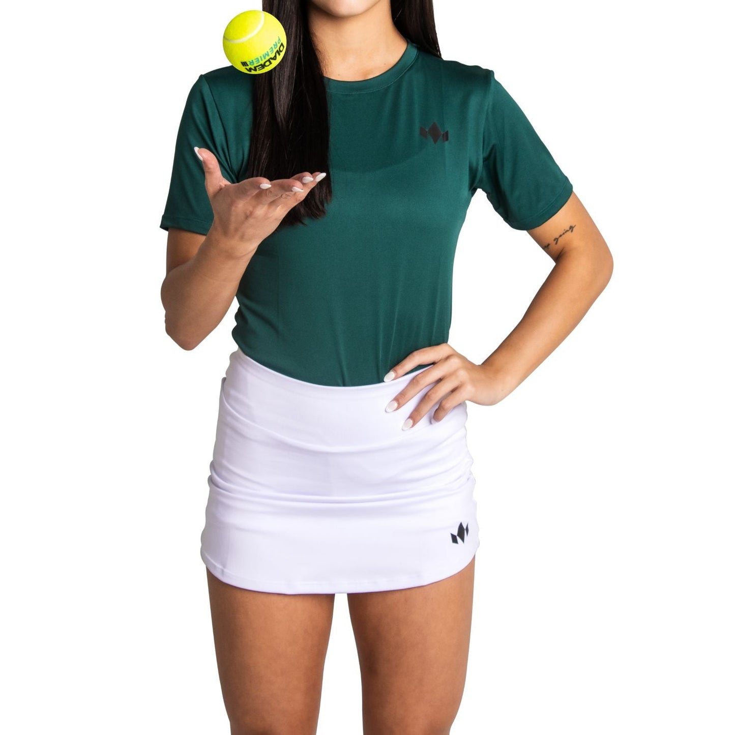 A woman with long dark hair, a forearm tattoo, and a hidden face stands against a white background wearing the Diadem Sports Women's Essential Tennis Skirt in fast-drying fabric and a green T-shirt, tossing a yellow tennis ball with one hand on her hip.