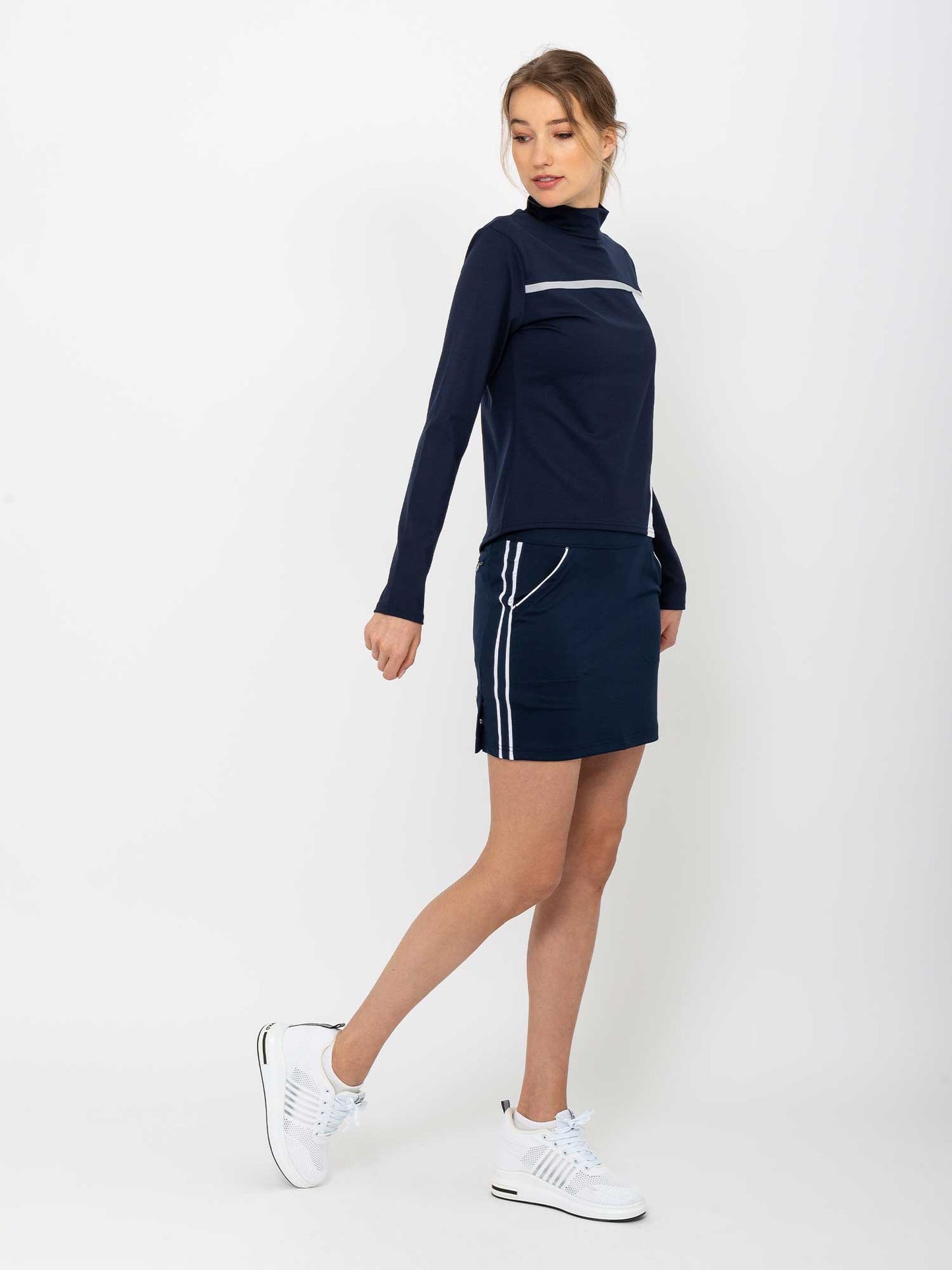 A woman in a navy long-sleeve top and the inPhorm NYC Women's Golf 15" Eloise Skirt in Midnight/White stands against a white background, wearing white sneakers. Her hair is tied back as she looks to the side, stepping forward.