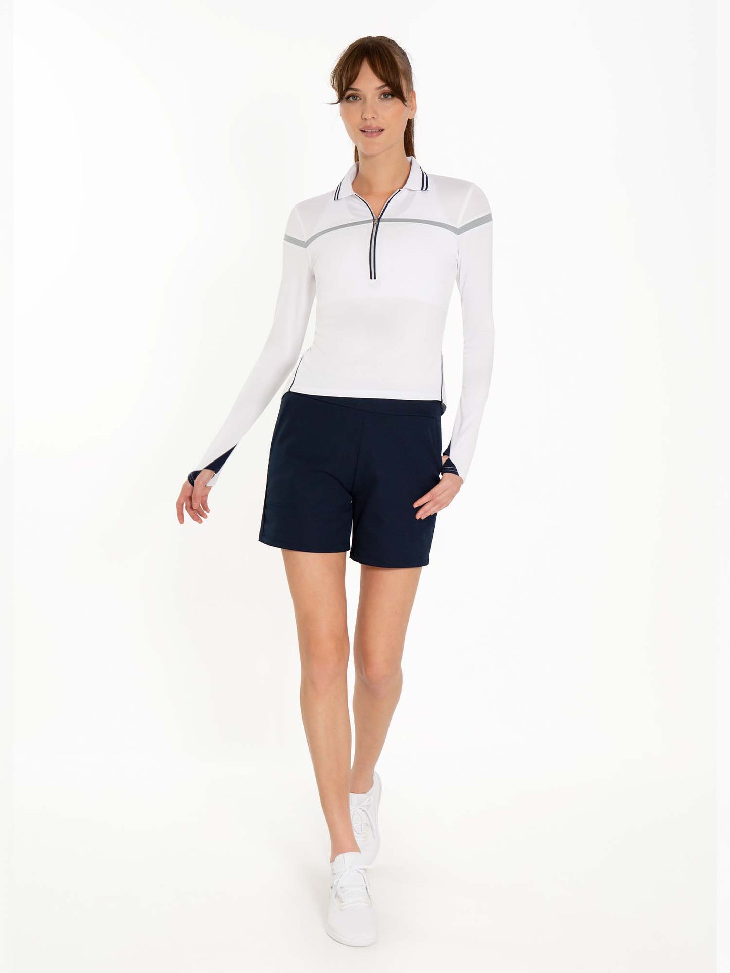 A woman with light skin and brown hair in a ponytail wears white sneakers, a white long-sleeve zip-up top with dark trim, and inPhorm NYC Women's Golf Shorts - Midnight against a white background. Her left arm is relaxed as she steps forward.