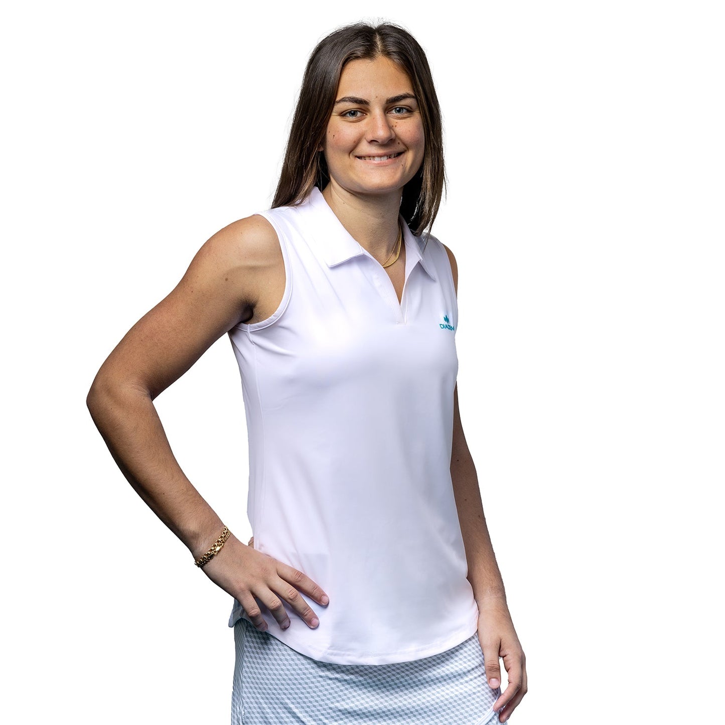 A woman with straight brown hair smiles gently, standing with one hand on her hip. She wears the Diadem Sports Women's Scales Bahama Sleeveless Polo and a light blue textured skirt against a plain white background.