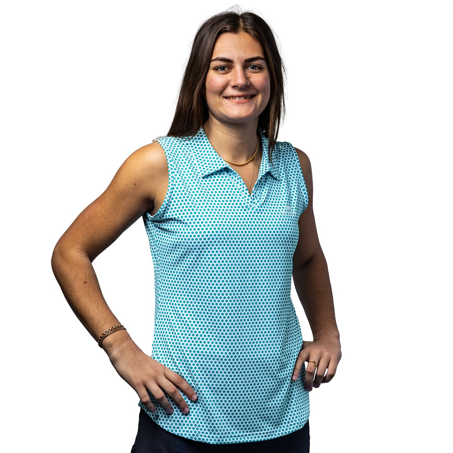 A woman with long brown hair smiles at the camera, wearing the Diadem Sports Women's Scales Pickleball Sleeveless Polo with UV 50+ protection and a dark blue dotted pattern, paired with dark shorts, standing confidently against a white background.