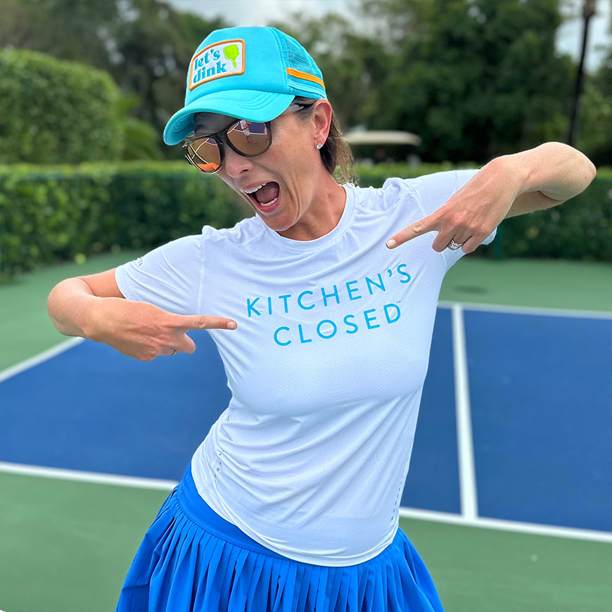 On the court, a woman proudly points to her Swinton Pickleball Women's Lux Performance White Kitchen's Closed Tee. She stays cool in the moisture-wicking shirt, styled with a blue skirt, reflective sunglasses, and a turquoise hat.