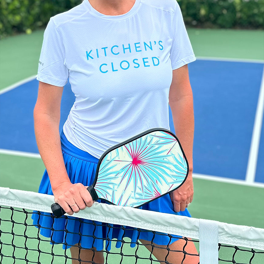 Standing behind the net on a pickleball court, she wears the Swinton Pickleball Women's Lux Performance White Kitchen's Closed Tee and a blue pleated skirt, holding a colorful paddle with a pink and teal burst design.
