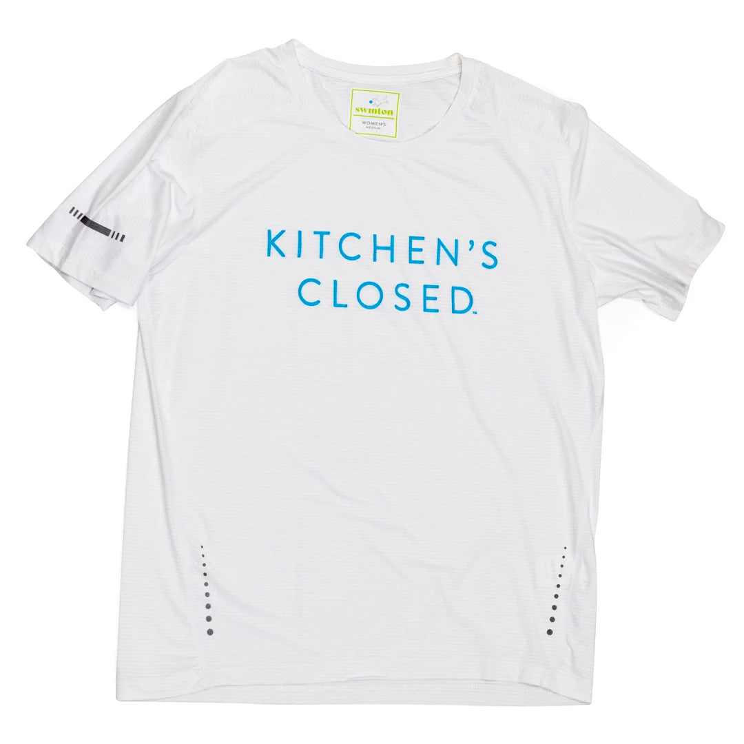 The Swinton Pickleball Women's Lux Performance White Kitchen's Closed Tee features blue “KITCHEN’S CLOSED” text, small black dot details near the hem, a visible inside neck label, and a sleeve logo—perfect for pickleball.