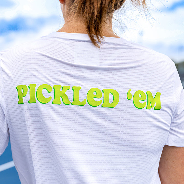 A woman with light brown hair in a ponytail is seen from behind wearing the Swinton Pickleball Women's Pickled 'em LUX Performance Shirt. The white shirt features bright green “PICKLED ’EM” text. A blue sports court and clouds are visible in the background.