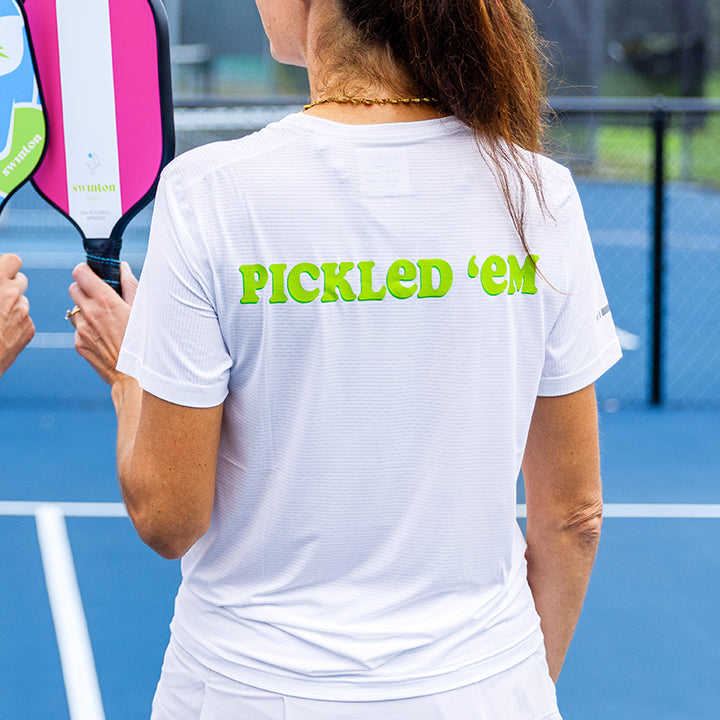 A woman on a blue pickleball court wears the Swinton Pickleball Women’s Pickled ’em LUX Performance Shirt with “PICKLED ’EM” in green on the back, holding a paddle. Another player stands nearby by the net and chain-link fence.