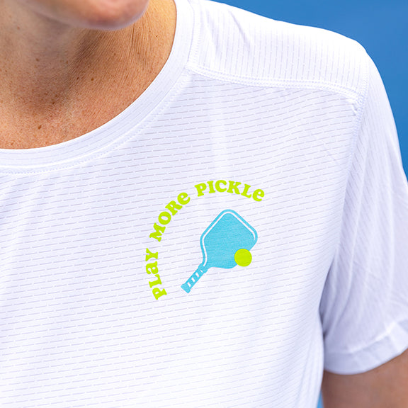 A woman wears the Swinton Pickleball Women's Pickled 'em LUX Performance Shirt, featuring a blue paddle, yellow ball, and "PLAY MORE PICKLE" text. The white shirt is shown on her upper torso and part of her chin.