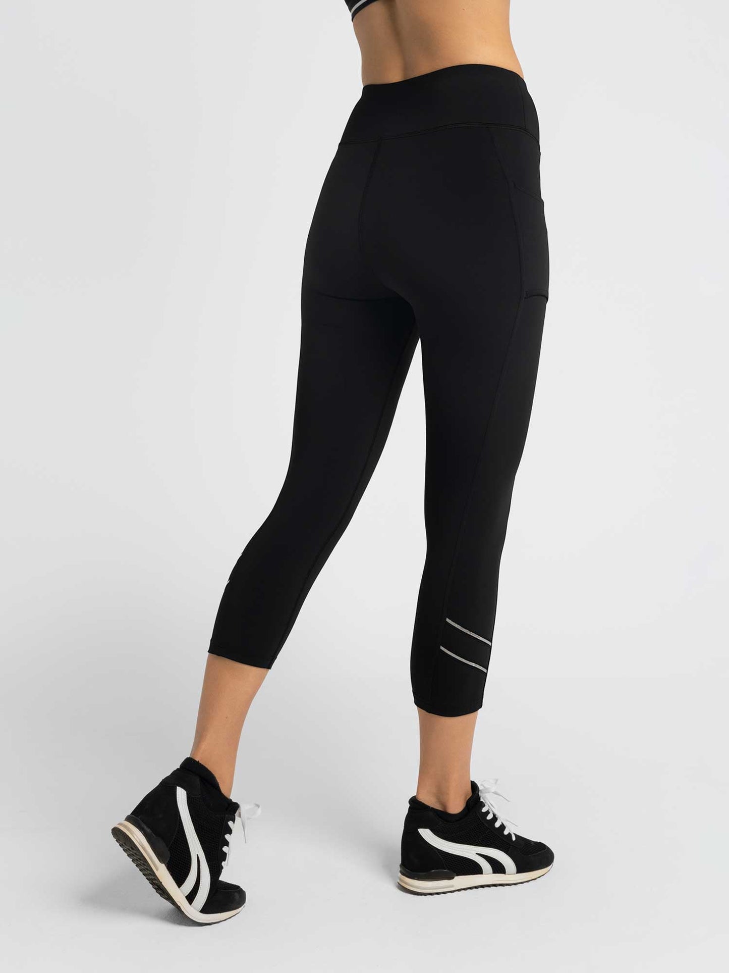 A person models the inPhorm NYC Zone Capri Legging - Black/Silver, featuring high waist, moisture-wicking fabric, reflective calf stripes, and a side pocket. Their back and legs are visible against a white background.