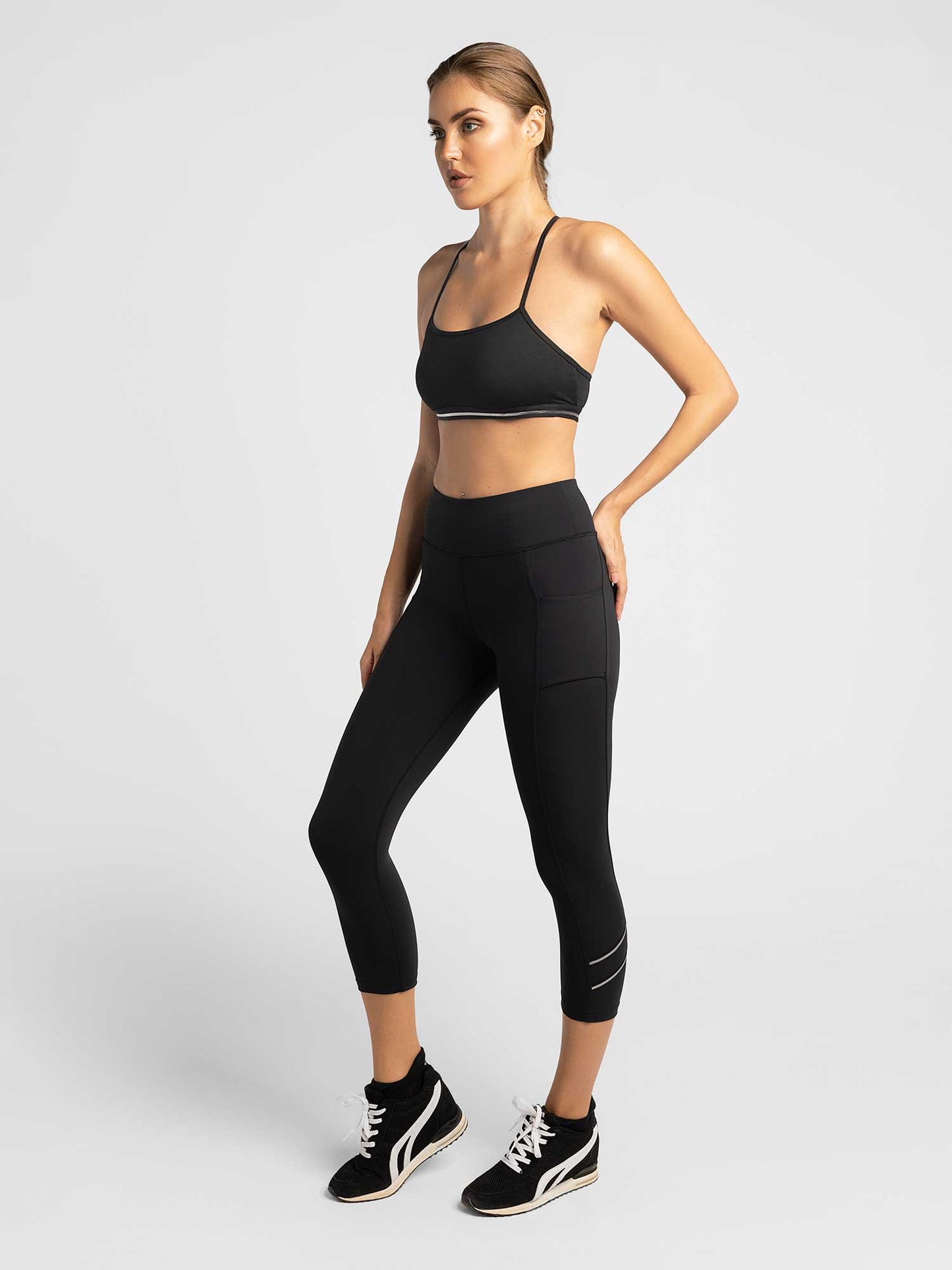 A woman poses in a studio against a light background, wearing inPhorm NYC’s Zone Capri Legging in Black/Silver with side pockets, a black sports bra, and black athletic sneakers. Her hair is tied back and she stands relaxed.