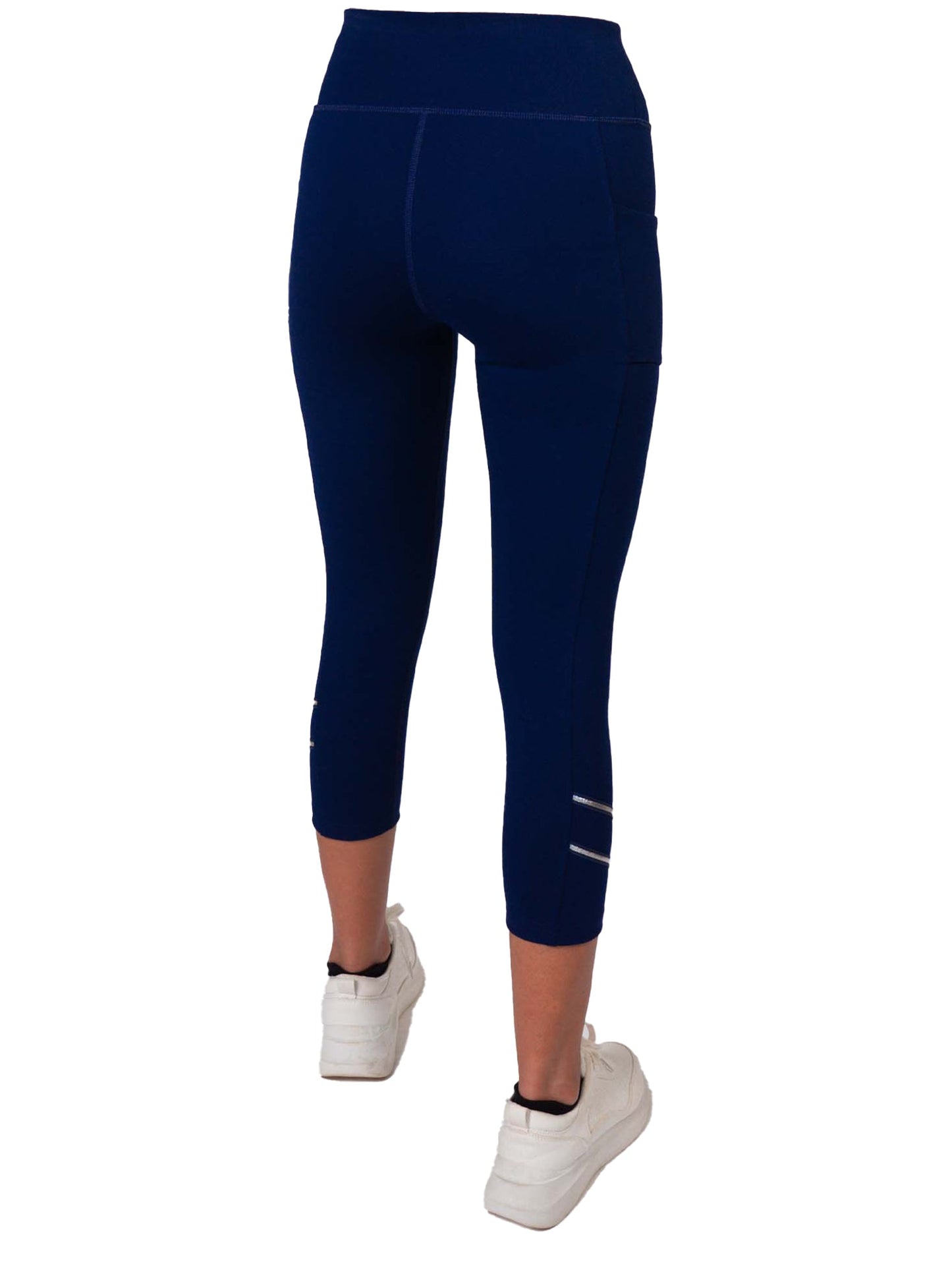 A person is shown from the back wearing inPhorm NYC’s Zone Capri Legging in Ink/Silver, featuring high-waisted compression, side seams, reflective calf stripes, and paired with white sneakers against a plain white background.