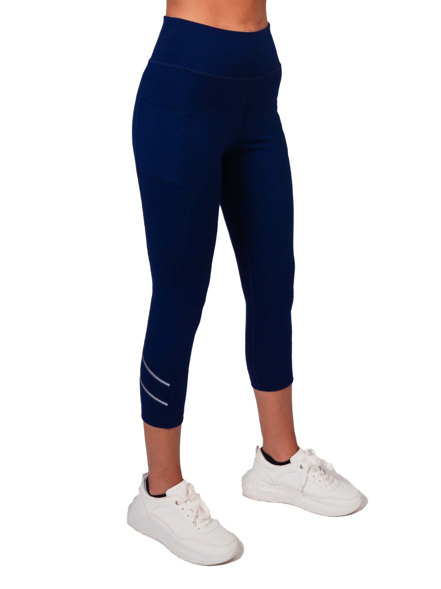 A person wears the inPhorm NYC Zone Capri Legging - Ink/Silver, featuring side pockets, a high-waisted fit, and two reflective stripes near the left calf, paired with white sneakers against a white background. Upper body mostly out of frame.