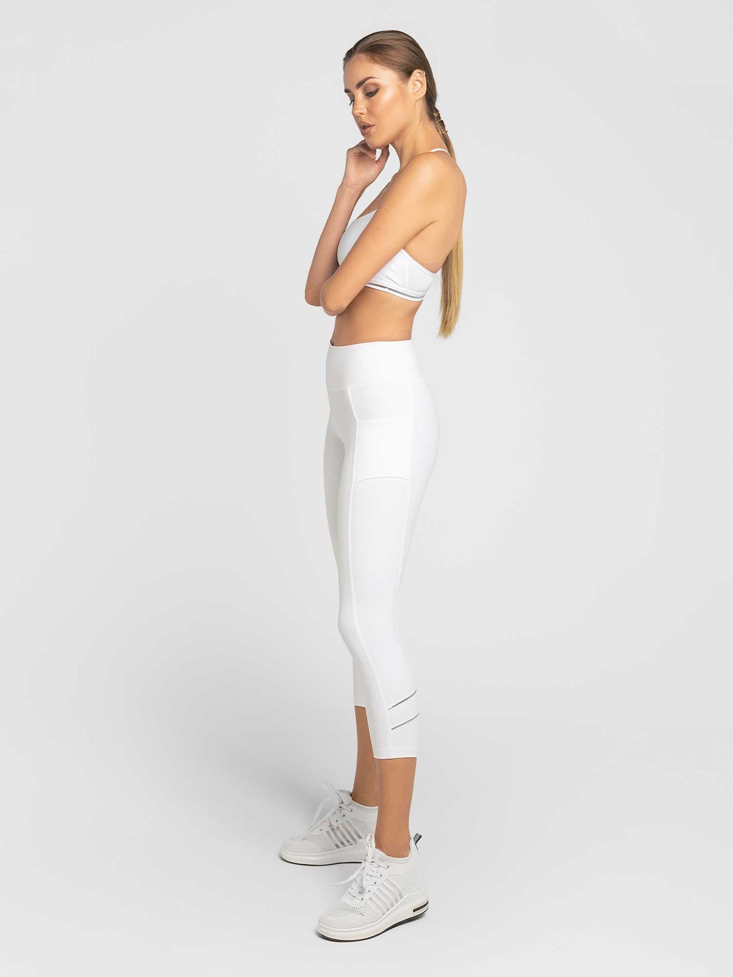 A woman with long, straight hair in a low ponytail stands in profile against a white background, thoughtfully resting her chin on her hand. She wears the inPhorm NYC Zone Capri Legging in White/Silver and a white sports bra with white sneakers.