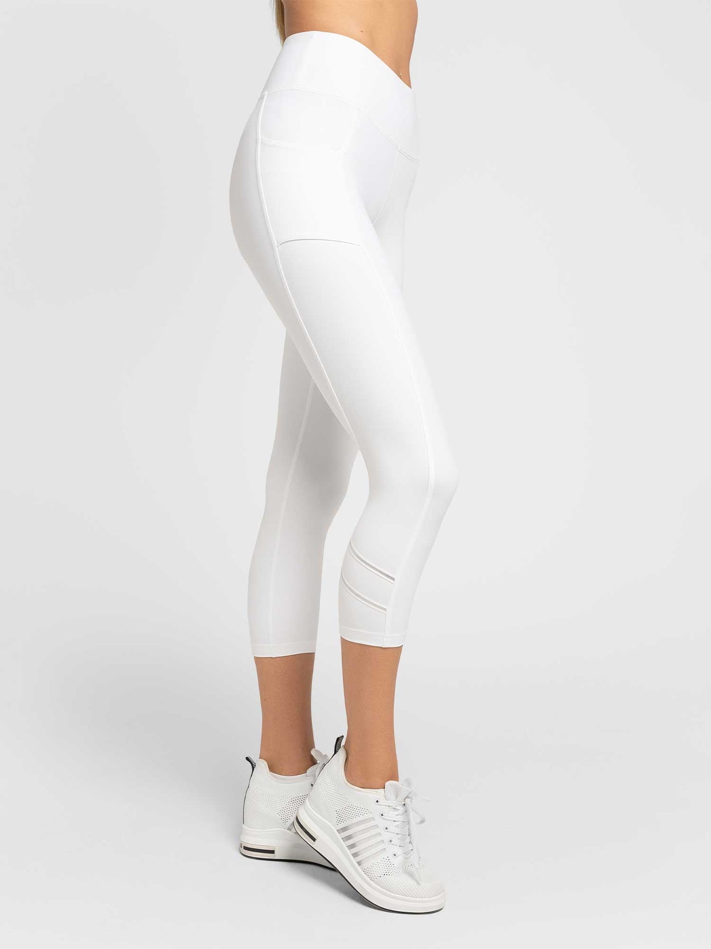 A person models the inPhorm NYC Zone Capri Legging in White/Silver, showing its high-waisted, moisture-wicking fit and side pocket. Paired with white sneakers, they stand sideways against a light gray studio backdrop; only their lower body is visible.