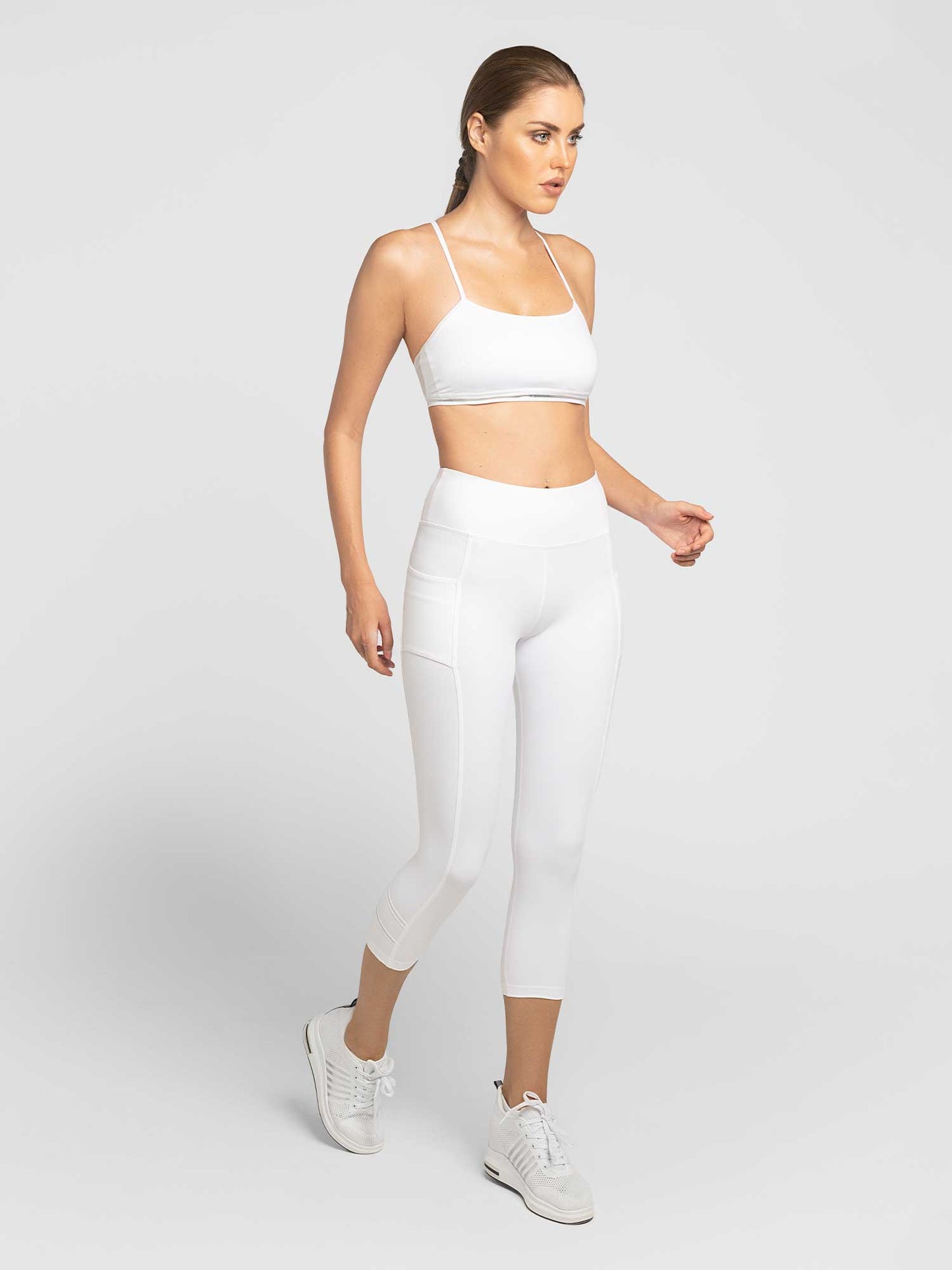 A woman with long brown hair tied back models the inPhorm NYC Zone Capri Legging in White/Silver with a white sports bra and sneakers, posing against a plain light gray background and looking slightly left with a neutral expression.
