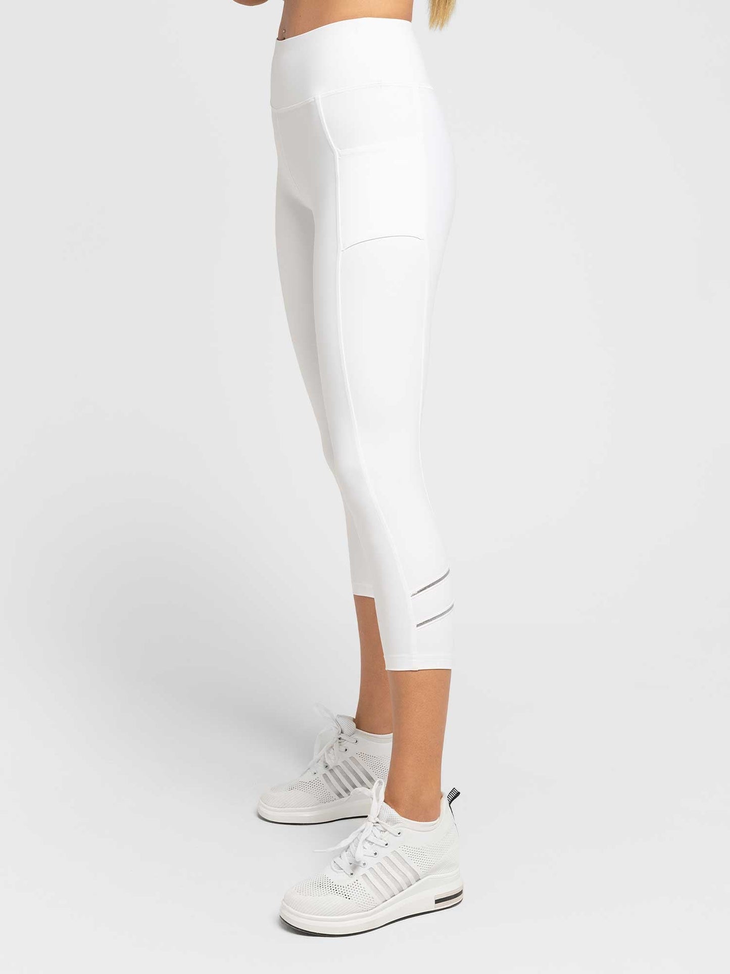 A person models the inPhorm NYC Zone Capri Legging - White/Silver, featuring high-waist compression, side pocket, moisture-wicking fabric, and mesh calf detail, paired with white sneakers against a plain white background. Only the lower body is visible.