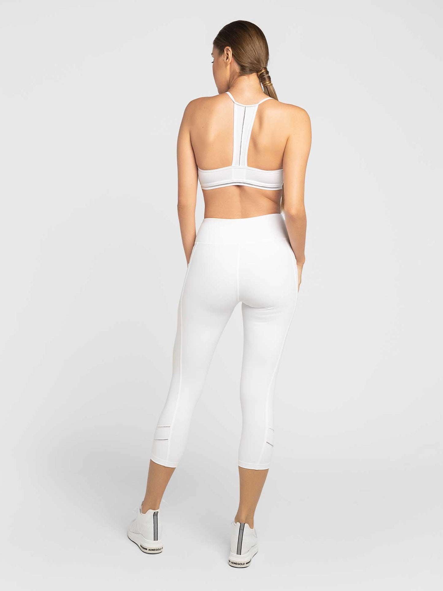 A woman with braided hair faces away, wearing the inPhorm NYC Zone Capri Legging - White/Silver and a white racerback sports bra, paired with white sneakers, set against a plain light gray background.